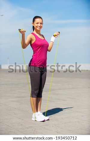 Ready to jump. Full length of beautiful young woman in sports clothing holding jumping rope and smiling while standing outdoors