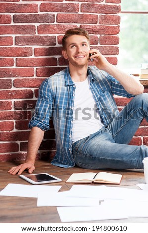 Good talk. Handsome young man sitting on the floor and talking on the mobile phone while digital tablet and documents laying near him