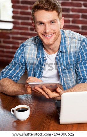 Examining his new tablet. Top view of handsome young man working on digital tablet and smiling while sitting at the table