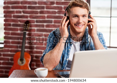 Enjoying music. Handsome young man in headphones working on laptop and smiling while acoustic guitar laying in the background
