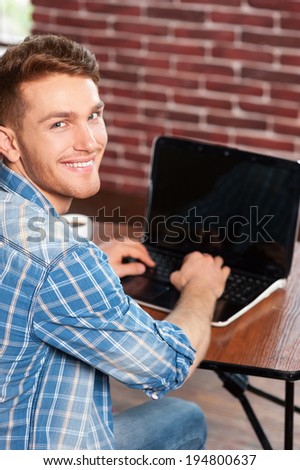 Surfing the net is fun. Top view of handsome young man working on laptop and looking over shoulder while sitting at his working place