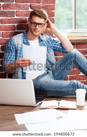 Man working at home. Handsome young man sitting on the floor and examining document while laptop and documents laying near him
