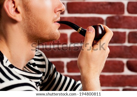 Smoking a pipe. Close-up of young man in striped clothing smoking pipe while sitting on the chair
