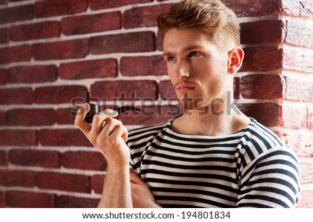 Sailor man. Handsome young man in striped shirt smoking pipe and looking away while leaning at the brick wall