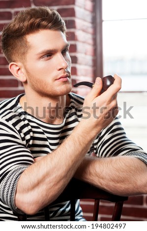 Smoking pipe. Handsome young man in striped shirt smoking pipe and looking away while sitting on the chair