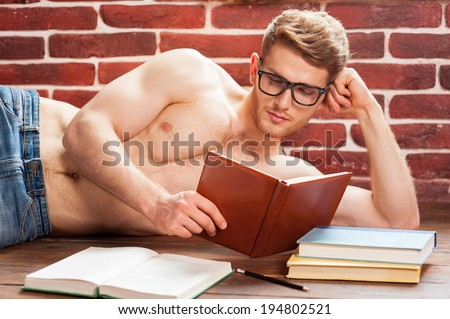Reading his favorite book. Thoughtful young shirtless man reading book while lying on the floor at his apartment