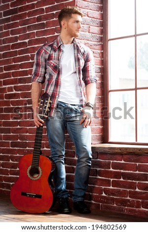In search of inspiration. Full length of handsome young man holding acoustic guitar and looking through window