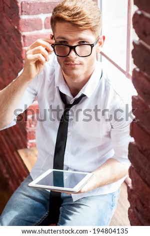 Using opportunities of digital age. Top view of handsome young man in shirt and tie working on digital tablet and looking at camera while sitting at the window sill