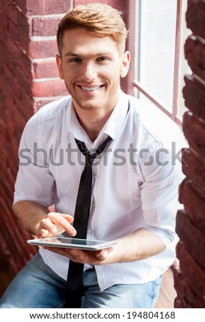 Testing his brand new tablet. Top view of handsome young man in shirt and tie working on digital tablet and smiling while sitting at the window sill