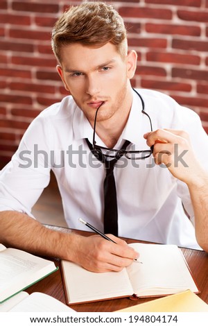 I need some fresh ideas. Top view of thoughtful young man in shirt and tie writing something in note pad and looking away while sitting at his working place