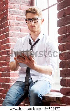 Handsome with digital tablet. Handsome young man in shirt and tie working on digital tablet and looking at camera while sitting at the window sill