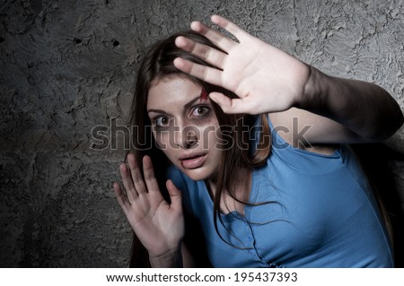 Please no! Terrified young woman looking at camera and stretching out hand while leaning at the dark wall