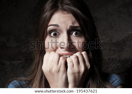 I am so scared! Shocked young woman staring at camera while holding fingers in mouth while standing against dark background