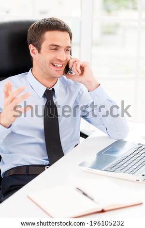 Happy businessman. Happy young man in formalwear looking at computer monitor and talking on the mobile phone while sitting at his working place