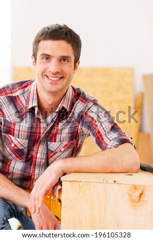 Handyman relaxing after work. Handsome young handyman looking at camera and smiling while sitting in workshop and leaning at the wooden deck