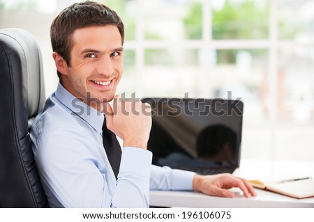 Confident and successful. Handsome young man in formalwear looking over shoulder and smiling while sitting at his working place