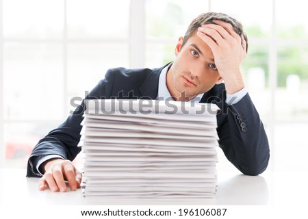 Missing deadlines. Depressed young man in shirt and tie looking at camera and holding head in hand while sitting at the table with stack of documents laying on it