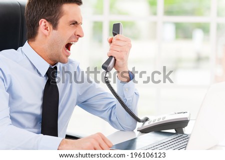I want this report now! Furious young man in shirt and tie shouting at the telephone while sitting at his working place