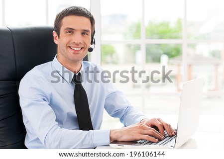 Cheerful male operator. Side view of cheerful young man in headset looking at camera and smiling while using laptop