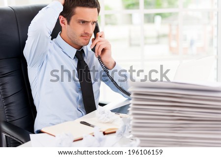 Busy office life. Frustrated young man in formalwear talking on the telephone and holding hand in hair while sitting at his working place