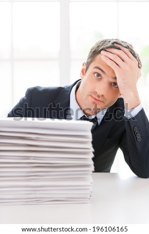 Depressed and overworked. Depressed young man in shirt and tie looking at camera and holding head in hand while sitting at the table with stack of documents laying on it