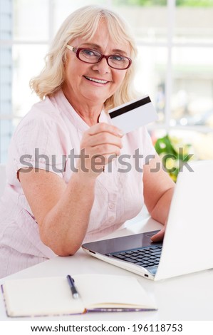 Shopping online. Happy senior woman using laptop and showing her credit card while sitting at the table