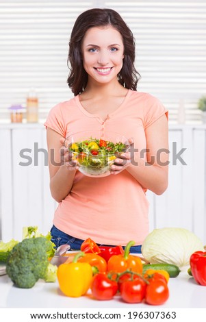 Only healthy food. Beautiful young woman holding a bowl with salad and looking at camera with smile while standing in the kitchen