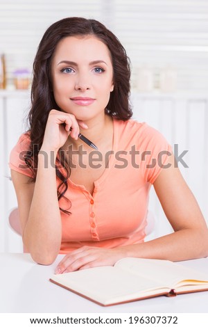 I need some fresh ideas. Beautiful young woman holding hand on chin and looking away while sitting at the table with note pad on it