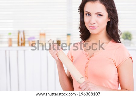 Housewife with rolling pin. Beautiful young woman holding rolling pin and looking at camera while standing in a kitchen