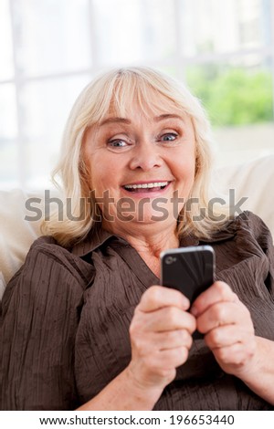 Senior woman with mobile phone. Cheerful senior woman holding mobile phone and smiling while sitting in a chair