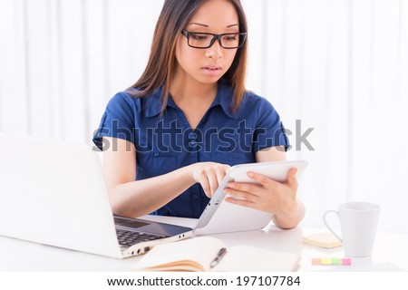 Using the advantages of digital age. Beautiful young Asian woman working on digital tablet while sitting at her working place