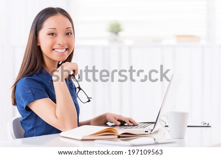 Young and confident. Attractive young Asian woman leaning her face on hands and smiling while sitting at her working place