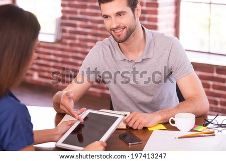 Let me show you all the features. Two cheerful people in casual wear sitting face to face at the table while man pointing digital tablet and smiling