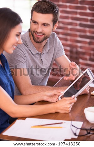 Discussing new business project. Side view of cheerful young man and woman sitting at the table and talking while man pointing digital tablet and smiling
