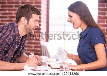 Battle of the sexes. Side view image of angry man and woman sitting face to face at the office table and shouting at each other