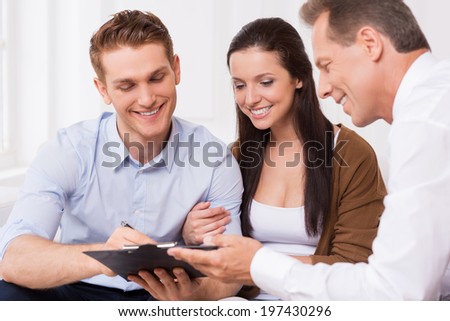 Just sign here. Happy young couple signing documents while confident mature man holding clipboard and smiling