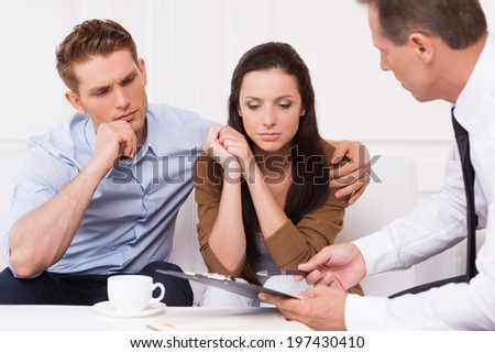 Expert advice. Thoughtful young couple sitting on the couch while confident financial advisor explaining something and pointing clipboard