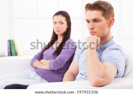 I wish we could work it out. Depressed young man holding hand on chin and looking away while angry woman sitting behind him on the couch and keeping arms crossed
