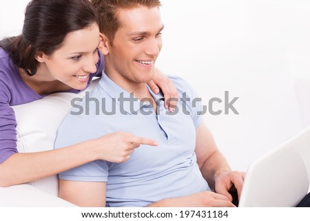 Can you believe it! Cheerful young loving couple sitting on the couch and looking at laptop while woman pointing monitor and smiling
