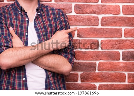 Pointing copy space. Close-up of man keeping arms crossed and pointing at copy space while standing against brick wall