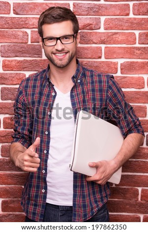 Confident IT guy. Handsome young man pointing you and smiling while holding laptop and standing against brick wall