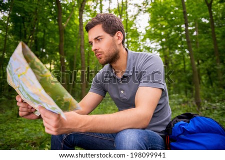 Where I am? Concentrated young man with examining map while sitting in a forest with backpack laying near him