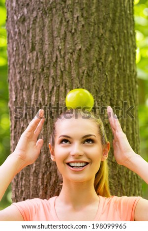 Carrying apple on head. Playful young woman carrying apple on head and looking up with smile while leaning at the tree in a park