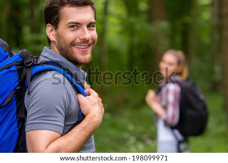 I love traveling. Handsome young man with backpack looking over shoulder and smiling while walking through a forest with woman in the background