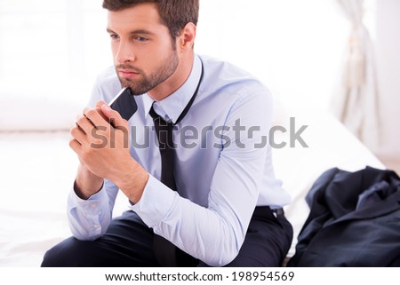 Lost in business thoughts. Thoughtful young man in shirt and tie touching his chin with mobile phone and looking away while sitting on the bed