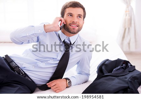 Executive on the go. Handsome young man in shirt and tie talking on the mobile phone and smiling while lying in bed