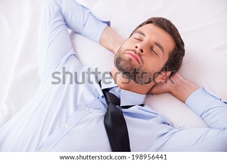 Businessman sleeping. Top view of handsome young man in shirt and tie holding hands behind head while sleeping in bed