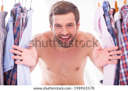 Shirts on every day. Happy young shirtless man looking through a various shirts hanging on hangers and smiling