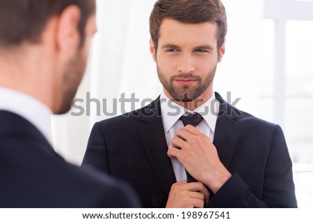 Looking just perfect. Handsome young man in formalwear adjusting his necktie and smiling while standing against mirror
