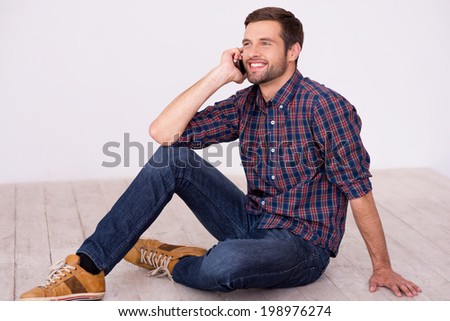 Good talk with friend. Handsome young man talking on the mobile phone and smiling at camera while sitting on hardwood floor